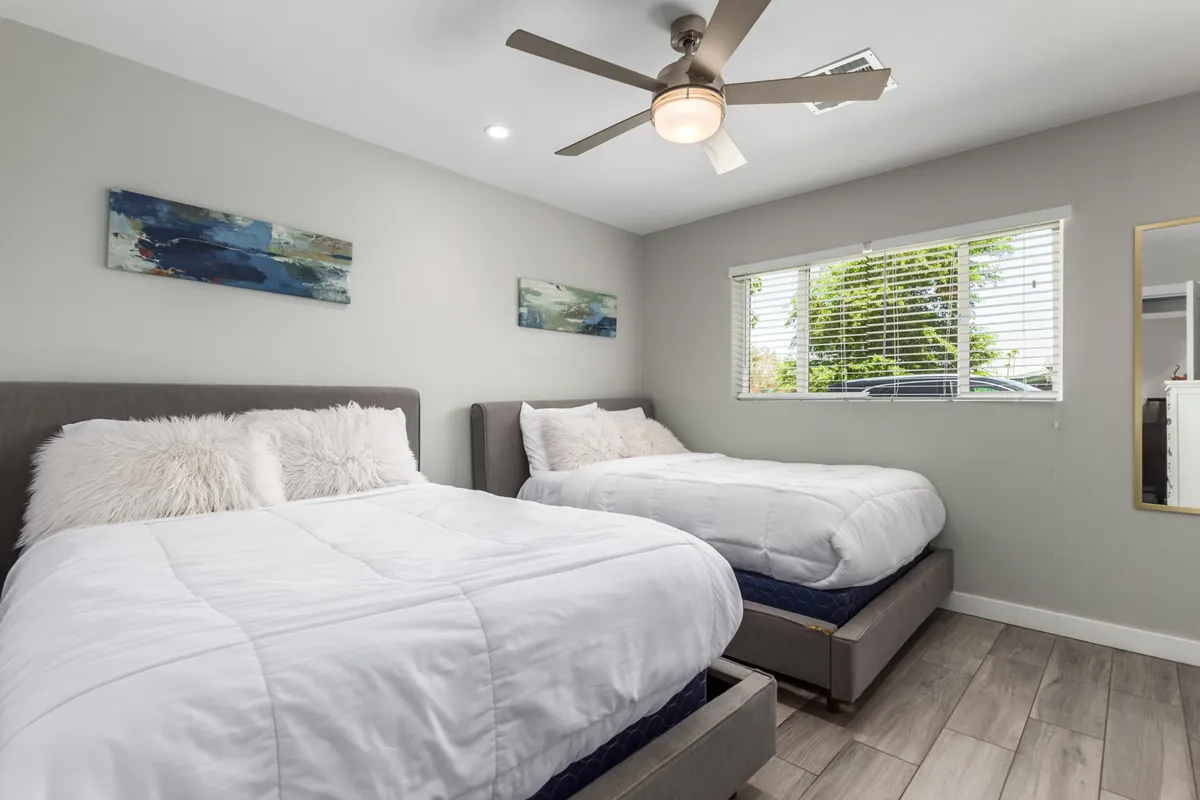 A spacious bedroom featuring two plush beds with crisp white bedding, decorative wall art, and a large window that bathes the space in natural light. A ceiling fan and modern furnishings create an airy, comfortable vibe, ideal for restful nights and group stays.