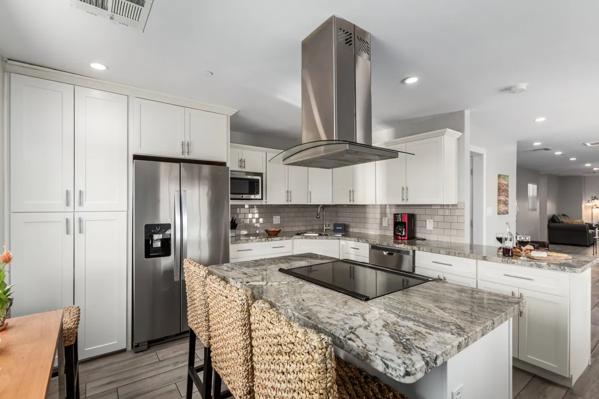 A spacious, well-appointed kitchen featuring a granite island countertop, stainless steel appliances, and woven bar stools at the breakfast bar. The layout is open and inviting, designed to encourage cooking, entertaining, and family gatherings, with ample prep space and modern finishes.
