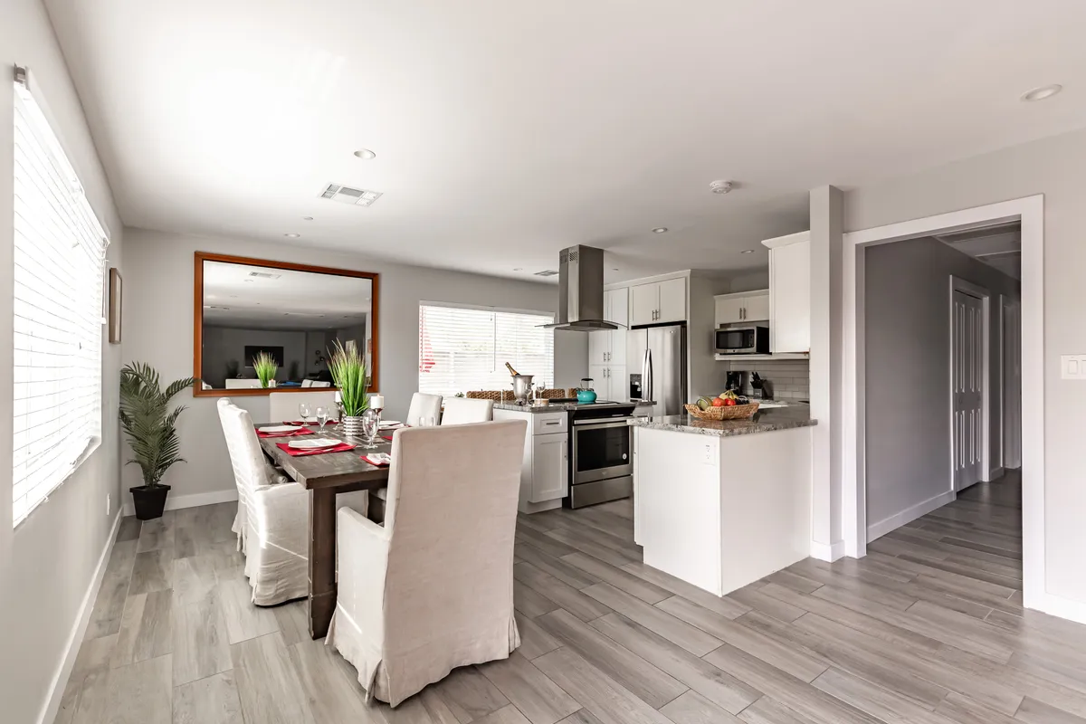 A view that seamlessly connects the dining area and kitchen, highlighting a dining table set for a group, the adjacent kitchen space, a decorative mirror, and a plant. The arrangement fosters an inviting environment for shared meals and conversations.