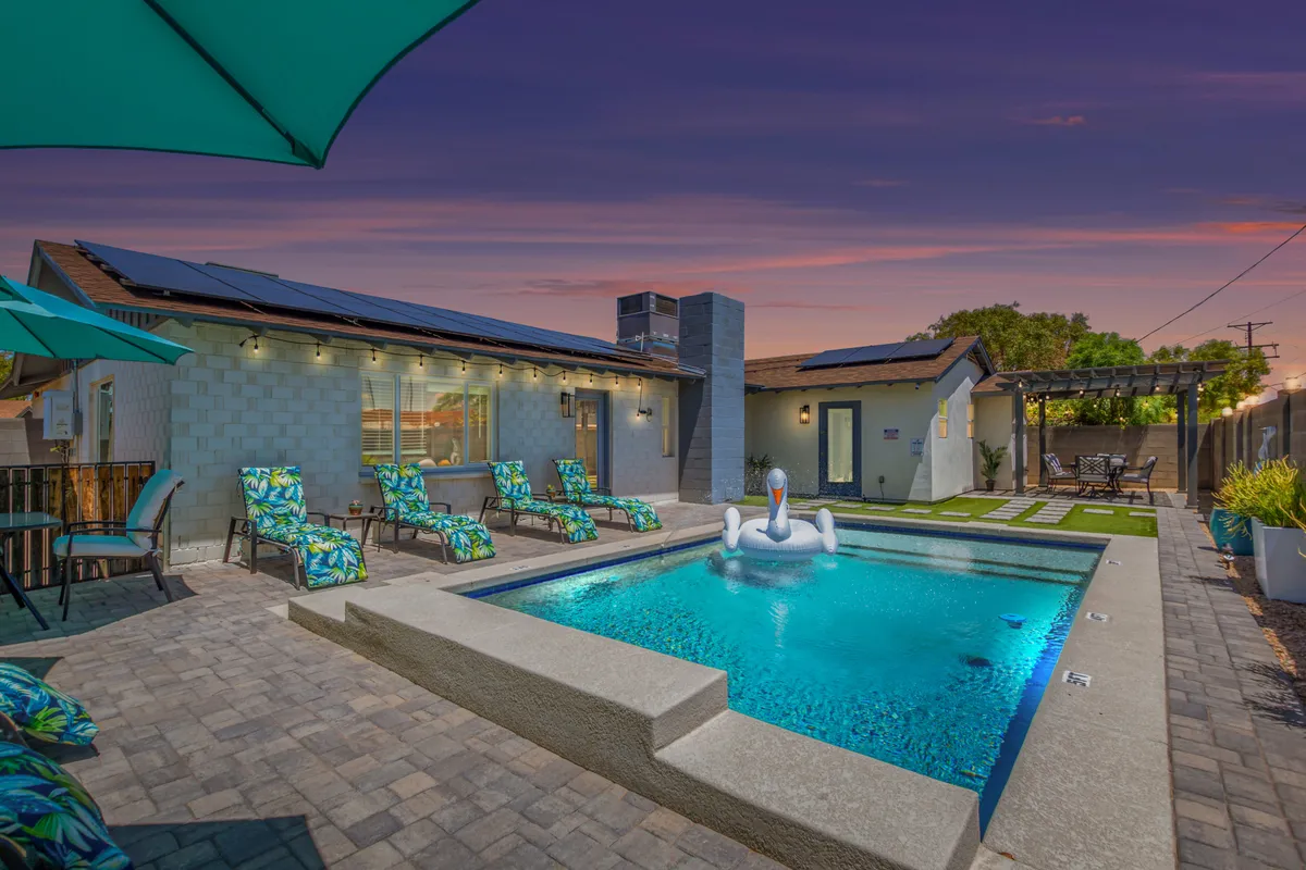 The image showcases a sunlit backyard oasis at sunset, featuring a sparkling swimming pool with a whimsical swan float, surrounded by chic lounge chairs and an inviting outdoor dining area. The setting hints at leisurely days swimming or sunbathing, and festive evenings gathering with friends or family under the Scottsdale sky.