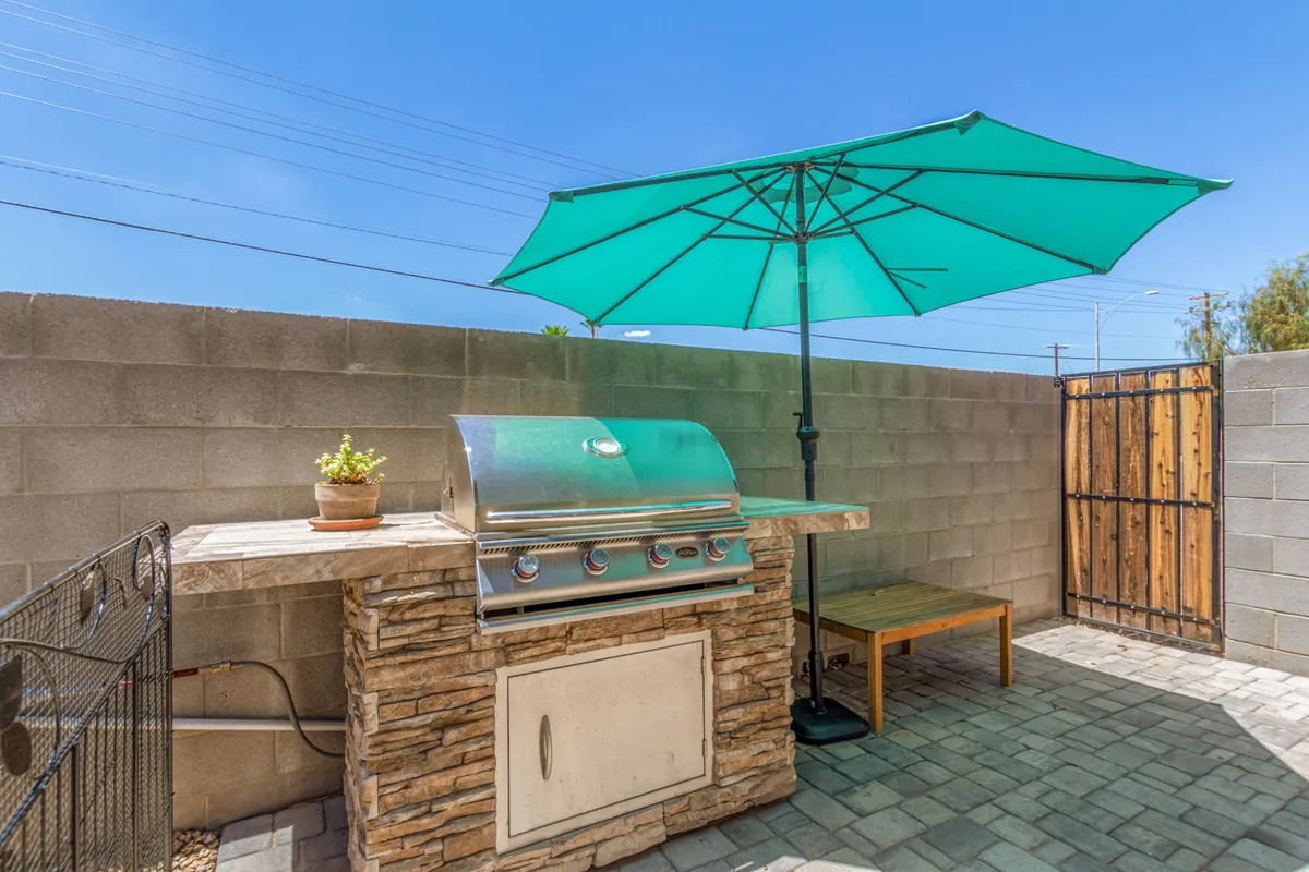 A paved outdoor area featuring a BBQ grill, umbrella, and a wooden gate in the background—suggesting both a dedicated grilling space and a secure, fenced environment for families and pets.