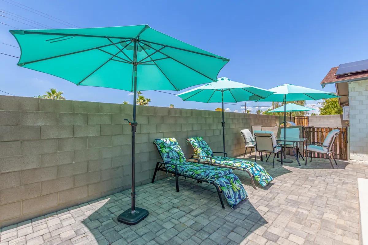 This exterior image features a backyard patio scene with turquoise umbrellas, lounge chairs adorned with tropical patterns, and a patio table with chairs. The setting is inviting and leisurely, suggesting opportunities for sunbathing, outdoor dining, and relaxing with friends or family.
