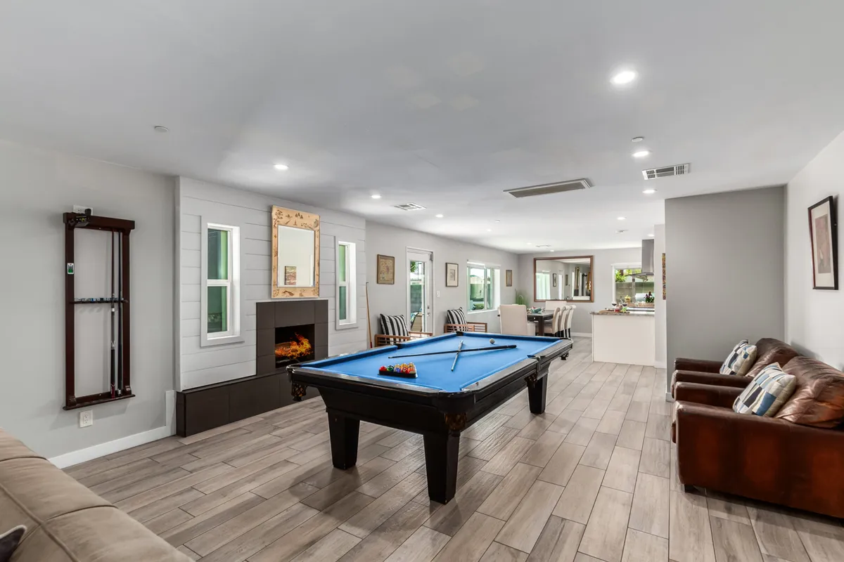 A vibrant open-plan living space with a pool table as a central feature, plush sofas arranged for conversation, a glowing indoor fireplace, and abundant natural light streaming through large windows.